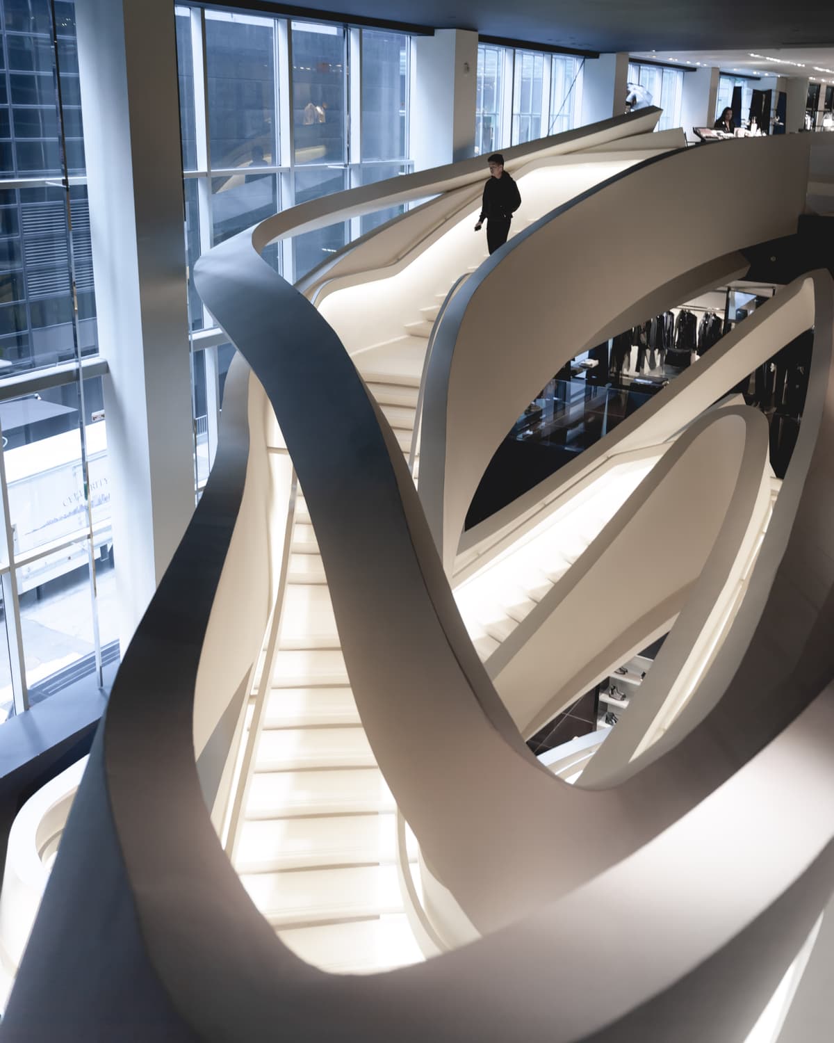 A sweeping sculptural white spiral staircase inside a luxury retail store, with a lone figure walking at the top