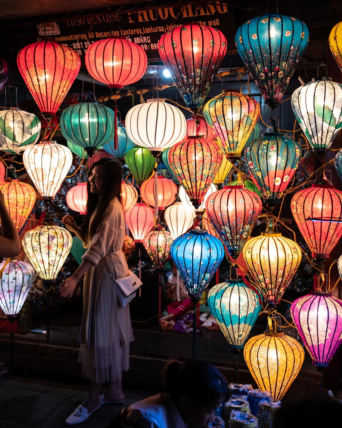 A woman stands among dozens of glowing colorful silk lanterns at a night market in Hoi An, Vietnam